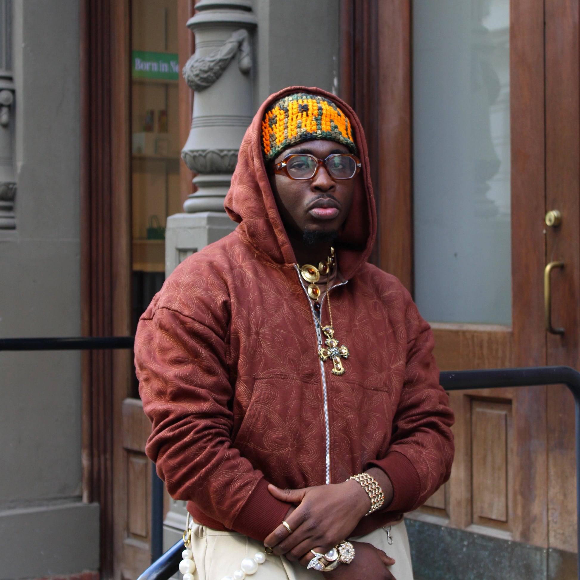 Man wearing a brown hoodie and glasses standing in front of a building entrance.